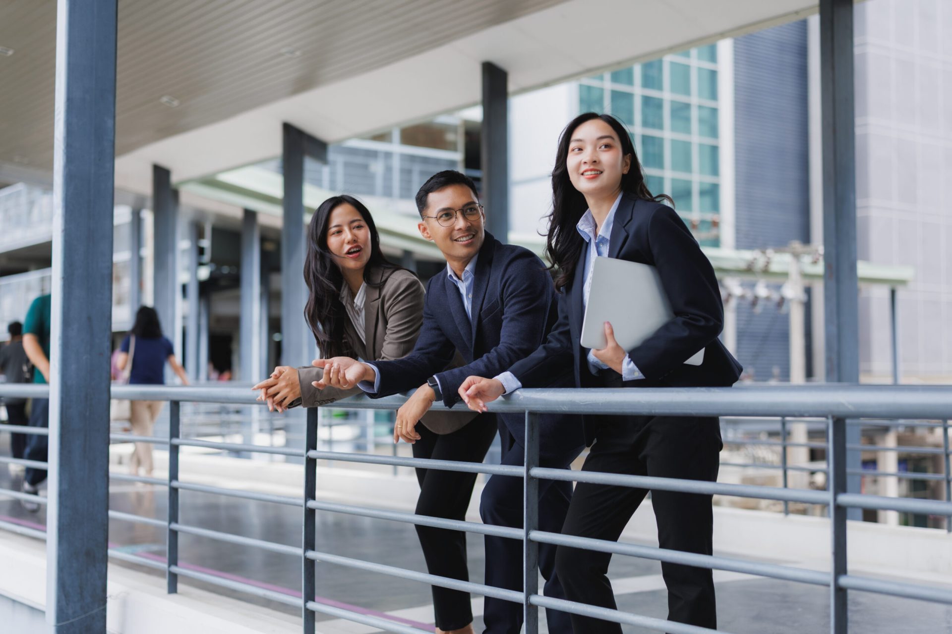 Three asian businesspeople are discussing a project, leaning on a railing in a modern office building